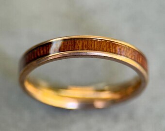 Puede incluir: Un anillo dorado con una incrustación de madera marrón oscuro. El anillo tiene una superficie pulida y reflectante, y la veta de la madera es visible. Es probable que sea una alianza de boda o un accesorio de moda.
