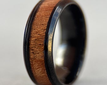 Op de afbeelding: Close-up van een ring met een donkere metalen band. Het midden van de ring heeft een band van lichtbruin hout, met zichtbare nerf. De ring is een eenvoudig, elegant ontwerp.