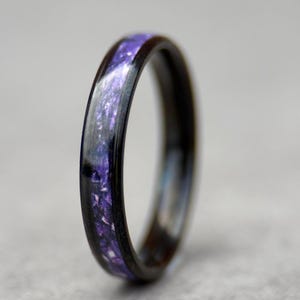 May include: A close-up of a ring with a dark band and a purple, speckled inlay. The ring's band appears to be made of a dark wood or metal, and the inlay has a shimmering, iridescent quality. The ring is set against a neutral background.