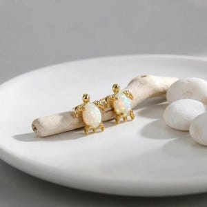 Pode incluir: Um par de brincos de tartaruga dourados com centros semelhantes a opalas. Os brincos são exibidos em um prato branco com um pedaço de madeira flutuante e pedras brancas. Os brincos têm um design detalhado.