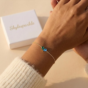 May include: A gold bracelet with green and blue gemstone charms is displayed on a wrist. A white box with the text "Skylosparkle" in gold script is in the background. The bracelet is delicate and elegant.
