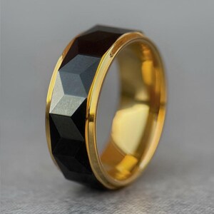 May include: A gold-coloured ring featuring a black, faceted band. The ring has a geometric design with a polished gold interior. The black band has a series of angled, triangular facets. The ring is a close-up shot against a grey background.