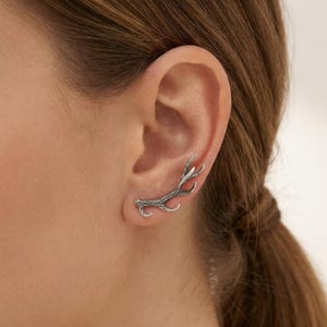 May include: A silver-toned ear cuff shaped like a deer antler, worn on the ear. The antler design has a textured surface and curves along the ear's edge, with multiple points extending upwards.