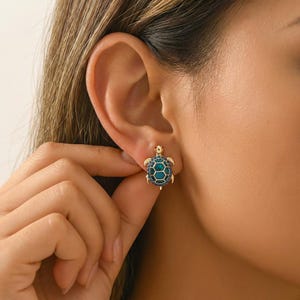 May include: Gold-toned turtle earrings with a teal and navy blue shell design. The earrings feature a detailed turtle shape with a textured shell and small red accents. The earrings are designed to be worn on the ear.