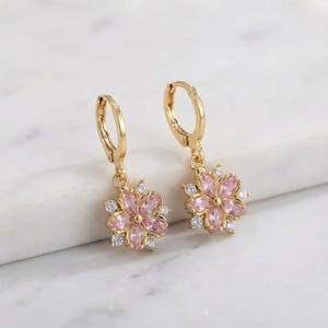 May include: A pair of gold-coloured hoop earrings, each with a pink flower pendant. The flower design is accented with small, clear gemstones. The earrings are displayed on a white surface.