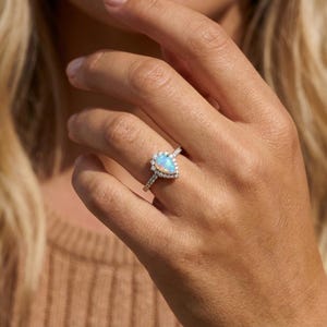 May include: A silver ring featuring a pear-shaped opal gemstone, surrounded by a halo of small, clear gemstones. The ring's band is also set with small, clear gemstones. The opal has a milky, iridescent appearance.