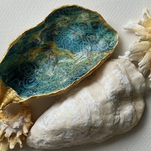 May include: An oyster shell dish with a gold-painted rim and an interior featuring a teal and blue swirling pattern. The exterior of the shell is off-white and textured, accompanied by two pale yellow, fan-shaped decorative coral pieces.