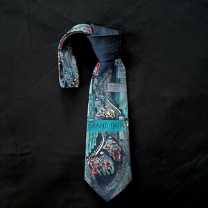 May include: A navy blue tie with a Grand Prix racing theme. The tie features a design with race cars, the words "Grand Prix", and other text. The tie is made of a smooth fabric and has a classic shape.