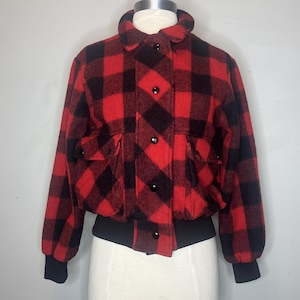 Pode incluir: Casaco bomber xadrez vermelho e preto com gola, punhos e cós pretos. O casaco tem dois bolsos frontais e botões pretos na frente. O casaco é exibido em um manequim.