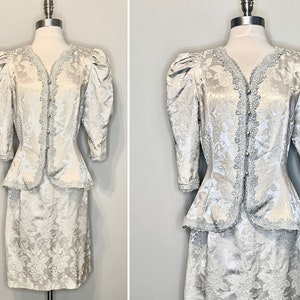 May include: A white and silver brocade skirt suit with a fitted jacket and a skirt. The jacket has a lace trim and a peplum waist. The skirt is knee-length and has a straight silhouette.