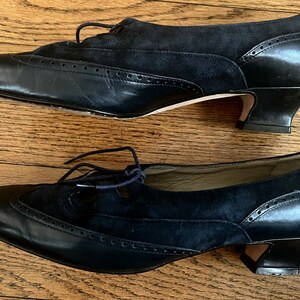 Puede incluir: Un par de zapatos de tac&oacute;n de cuero negro y ante con cierre de cordones. Los zapatos tienen punta puntiaguda y un dise&ntilde;o de brogue decorativo.