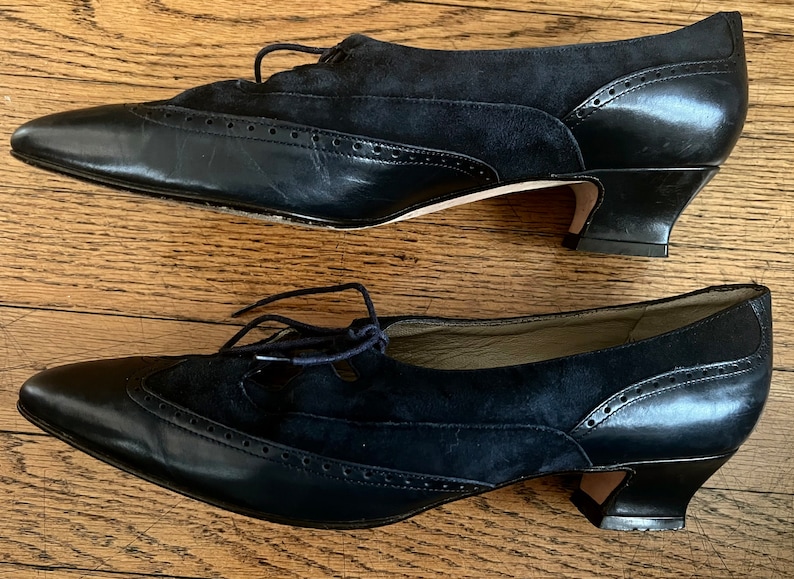 Puede incluir: Un par de zapatos de tac&oacute;n de cuero negro y ante con cierre de cordones. Los zapatos tienen punta puntiaguda y un dise&ntilde;o de brogue decorativo.