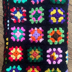 May include: A colourful, handmade crochet blanket. It features a black background with square floral patterns in pink, green, yellow, orange, and purple. The blanket has a red border. The blanket is made with yarn.