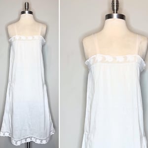 May include: A white sleeveless dress featuring thin shoulder straps and a square neckline. The garment is adorned with a delicate white floral embroidery pattern along the upper chest area and a matching embroidered border at the hemline.