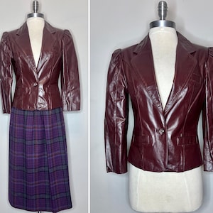 May include: A burgundy leather blazer with a single button closure and a tailored fit. The blazer is paired with a purple plaid skirt. The outfit is displayed on a mannequin.