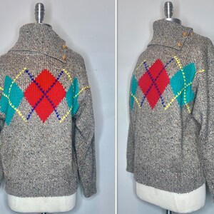 Könnte beinhalten: Ein grauer Strickpullover mit hohem Kragen und Argyle-Muster. Das Argyle-Design hat eine rote Raute in der Mitte, flankiert von türkisfarbenen Rauten, mit gelben und blauen Akzenten. Der Kragen hat zwei braune Knöpfe.