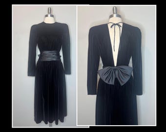May include: Two black velvet dresses on mannequins. The first dress has long sleeves and a wide satin belt. The second dress has a plunging neckline, long sleeves, and a large satin bow at the waist. Both dresses are knee-length.