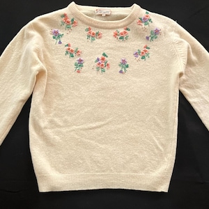 May include: A cream-colored sweater with a floral embroidered design around the neckline. The flowers are pink, purple, and green.