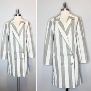 Op de afbeelding: Een double-breasted blazer met een verticaal gestreept patroon in wit en lichtgrijs. Het kledingstuk heeft lange mouwen, een reverskraag en twee rijen knopen aan de voorzijde, samen met twee grote opgestikte zakken op heuphoogte.