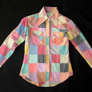 May include: A colorful patchwork shirt with long sleeves, a collar, and two chest pockets. The shirt features a button-down front and is composed of various pastel-colored squares, creating a unique and eye-catching design. The shirt is displayed on a black background.