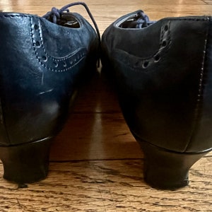 Puede incluir: Un par de zapatos de tac&oacute;n de cuero azul oscuro con dise&ntilde;o de brogue. Los zapatos tienen la punta cerrada y un cierre de cordones en la parte delantera.