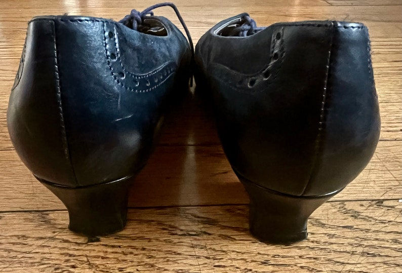 Puede incluir: Un par de zapatos de tac&oacute;n de cuero azul oscuro con dise&ntilde;o de brogue. Los zapatos tienen la punta cerrada y un cierre de cordones en la parte delantera.