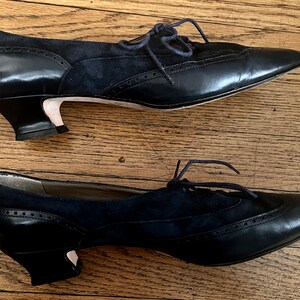 Puede incluir: Un par de zapatos de tac&oacute;n de cuero negro y ante con cordones. Los zapatos tienen una punta puntiaguda y un tac&oacute;n de bloque bajo.