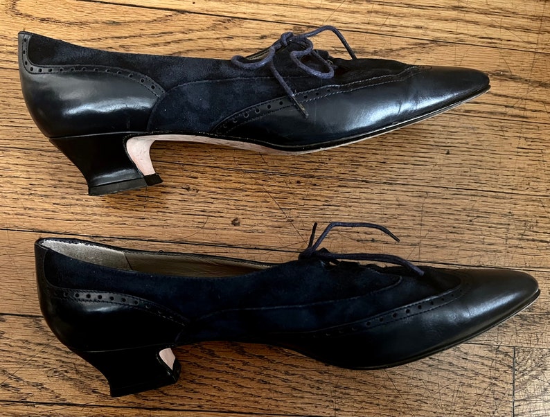 Puede incluir: Un par de zapatos de tac&oacute;n de cuero negro y ante con cordones. Los zapatos tienen una punta puntiaguda y un tac&oacute;n de bloque bajo.