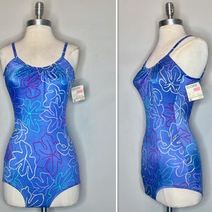 May include: A blue one-piece swimsuit with a floral pattern in white, purple, and light blue. The swimsuit has thin straps and a gathered neckline. The garment is displayed on a mannequin.