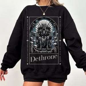 Puede incluir: Sudadera negra con un estampado gráfico de un esqueleto sentado en un trono rodeado de otros esqueletos. El texto "Dethrone" está impreso debajo de la imagen. El texto "Getting out is not enough if no body else can come" está impreso en el lado izquierdo de la imagen. El texto "If he's home I've got a message for the man above" está impreso en el lado derecho de la imagen.