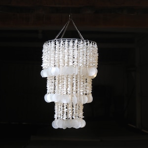 May include: A white shell chandelier with three tiers of hanging shells. The chandelier is made from natural materials and has a rustic, bohemian style.