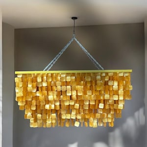 Op de afbeelding: Een gouden kroonluchter met een rechthoekig frame en een cascade-ontwerp van vierkante stukken. De kroonluchter hangt aan een ketting aan het plafond.