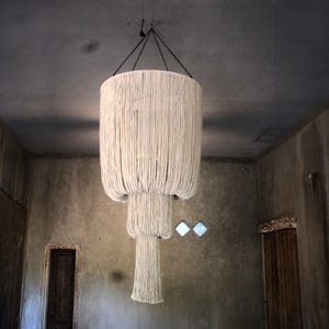 Op de afbeelding: Een grote, witte, geweven macramé-kroonluchter met meerdere lagen hangt aan het plafond in een kamer met een betonnen muur.