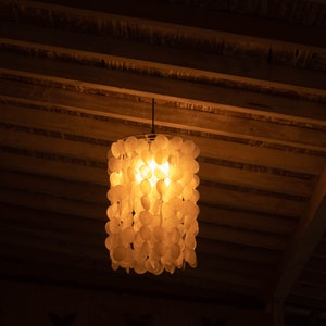 Op de afbeelding: Een witte schelp-chandelier met een warme gloeilamp die hangt aan een houten plafond.