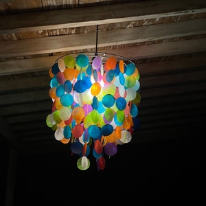 Puede incluir: Una lámpara colgante colorida y de múltiples capas hecha de conchas redondas, translúcidas e iridiscentes. La lámpara cuelga del techo y está iluminada, proyectando un brillo cálido.