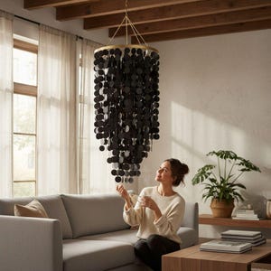 May include: A black chandelier with a gold-coloured metal frame and hanging circular accents. The chandelier is suspended from the ceiling in a living room setting. The room features a grey sofa, wooden beams, and a houseplant.