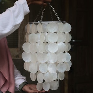 Könnte beinhalten: Ein weißer Muschel-Kronleuchter mit einem silbernen Metallrahmen. Der Kronleuchter besteht aus vielen einzelnen Muscheln, die in einem kaskadierenden Muster zusammengefügt sind.