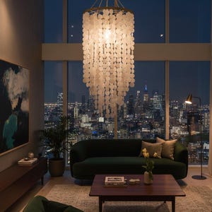 Puede incluir: Interior de un salón moderno de noche. Una gran lámpara de araña adornada con detalles circulares blancos cuelga del techo. Un sofá verde oscuro y un sillón a juego están frente a una gran ventana con vistas al horizonte de la ciudad.