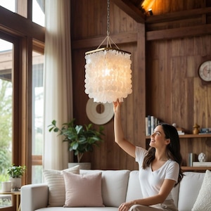 May include: A square pendant light fixture featuring multiple tiers of translucent, circular white capiz shell discs suspended from a metallic frame. The light is attached to a dark chain and emits a soft, warm glow from within the shell layers.