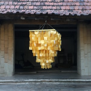Könnte beinhalten: Ein großer, quadratischer, goldfarbener Kronleuchter aus vielen kleinen, überlappenden, muschelförmigen Teilen. Der Kronleuchter hängt von der Decke eines Raumes mit einer Backsteinwand.