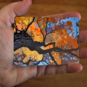 May include: A rectangular art print featuring a stylized tree with orange, yellow, and blue foliage against a light blue sky. The tree's dark, textured branches and trunk are prominent. The print is held in a hand.