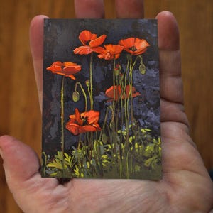 May include: A miniature painting of red poppies against a dark blue background. The artwork features several open blooms and buds with green stems and foliage. The painting is held in a hand, showcasing its small size.