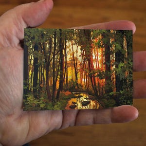 May include: A small rectangular painting depicts a forest scene with tall trees and a stream. The artwork features a warm color palette of orange, yellow, and green, suggesting a sunset or sunrise. The painting is held in a hand.