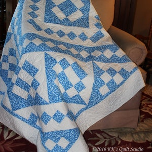 May include: A white and blue quilt with a diamond pattern. The quilt is folded and draped over a chair. The quilt is made with a blue and white fabric with a small floral pattern. The quilt is edged with a white border.