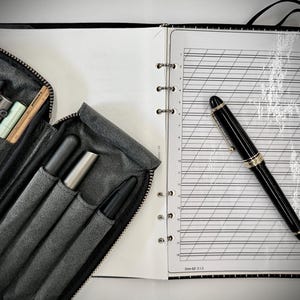 May include: A black fountain pen rests on an open ring-bound notebook with lined calligraphy paper. Beside it, a dark gray zippered pen case holds several writing instruments, including metallic and matte black pens and a wooden pencil.