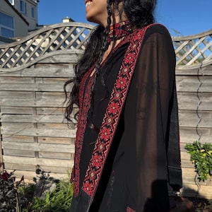 May include: Black tunic with red and white embroidered floral patterns on the sleeves and trim. The tunic has a matching scarf and shoulder strap. A brown leather bag is visible.
