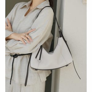 May include: A light gray leather shoulder bag with a curved design and black trim. The bag has a single shoulder strap and a silver-colored metal ring detail. The bag is held by a person wearing a light-colored linen outfit.