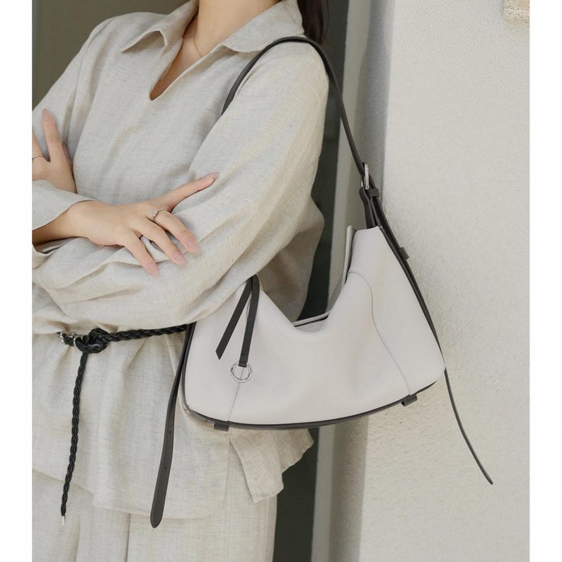 May include: A light gray leather shoulder bag with a curved design and black trim. The bag has a single shoulder strap and a silver-colored metal ring detail. The bag is held by a person wearing a light-colored linen outfit.