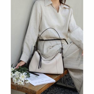 May include: Cream-colored leather handbag with dark brown accents. The bag has a curved shape, a top handle, and a silver-tone ring detail. The bag is sitting on a wooden surface next to a bouquet of white flowers and a magazine.