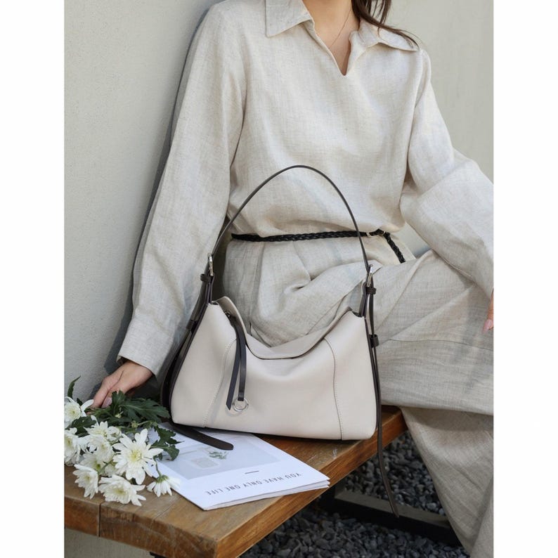 May include: Cream-colored leather handbag with dark brown accents. The bag has a curved shape, a top handle, and a silver-tone ring detail. The bag is sitting on a wooden surface next to a bouquet of white flowers and a magazine.